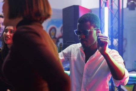 Photo for African american young man in sunglasses dancing in crowded nightclub while attending discotheque. Clubber partying on dancefloor with friends, celebrating and having fun in club - Royalty Free Image