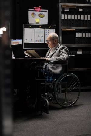 Photo for Senior wheelchair user checking annual profit revenue paperwork before business consulting meeting. Executive with paraplegia doing objective completion measurements in file cabinet office - Royalty Free Image