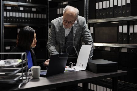 Photo for Business invoicing management employees comparing accountancy market data analysis between laptop and paperwork. Archival depository filled with document folders on cabinet shelves and graphic charts - Royalty Free Image