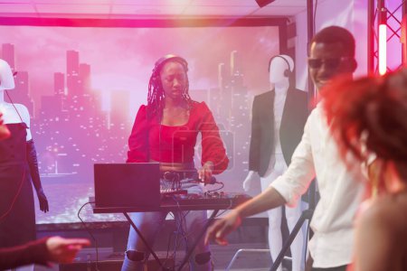 Téléchargez les photos : Femme DJ en utilisant la console de mixage pour mélanger les pistes sur scène à la soirée disco boîte de nuit. Musicien afro-américain dans les écouteurs en appuyant sur les boutons sur la station de musique électronique au club - en image libre de droit