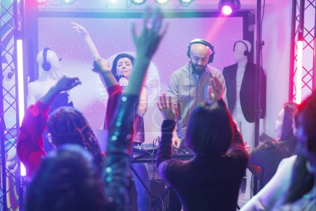 Crowd dancing and raising hands at band concert in nightclub. Singer and dj performing on stage with spotlights and engaging diverse people partying at discotheque on dancefloor