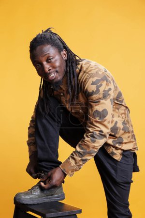 Photo for African american person tying shoelace on his stylish shoes, preparing for long walk. Man wearing fashionable clothes and sneakers standing in studio posing over yellow background. - Royalty Free Image