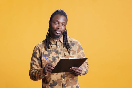 Photo for Young adult holding clipboard documentation, writing business job schedule on papers in studio over yellow background. African american man analyzing checklist, signing agreement files - Royalty Free Image
