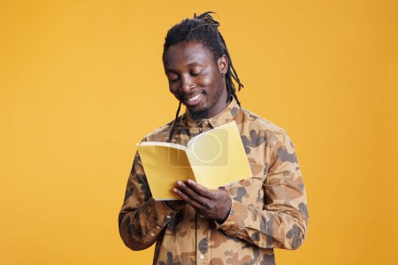Téléchargez les photos : Joyeux homme afro-américain lisant un livre de fiction, appréciant d'apprendre de nouvelles informations debout en studio sur fond jaune. Personne intelligente étudiant la littérature, aimer le genre et l'intrigue - en image libre de droit