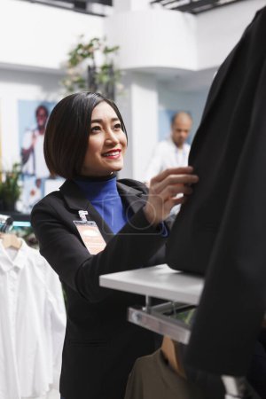 Téléchargez les photos : Magasin de vêtements souriant asiatique employé fermeture boutons de veste tout en portant mannequin dans des vêtements formels. Shopping centre commercial boutique travailleuse organisant des vêtements pour femmes à vendre - en image libre de droit