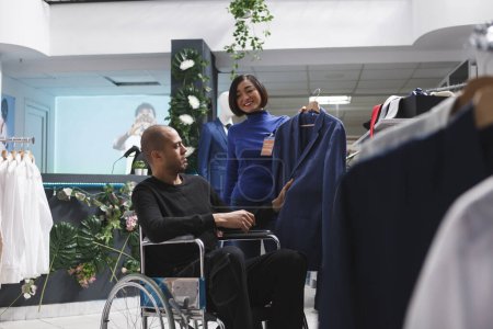 Photo for Clothing store asian woman worker consulting arab man customer with physical disability in choosing formal jacket. Boutique seller offering stylish outfit while assisting client in wheelchair - Royalty Free Image