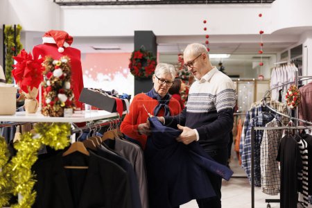 Photo for Senior clients looking for jackets on hangers, buying clothes on sale from clothing store at mall. Elderly people checking fashion items, searching formal attire and presents for christmas. - Royalty Free Image