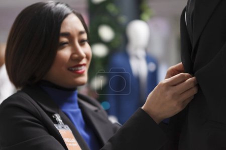 Photo for Clothing store smiling asian woman worker putting formal jacket on mannequin while managing inventory. Boutique cheerful seller fastening buttons and adjusting suit on dummy model - Royalty Free Image