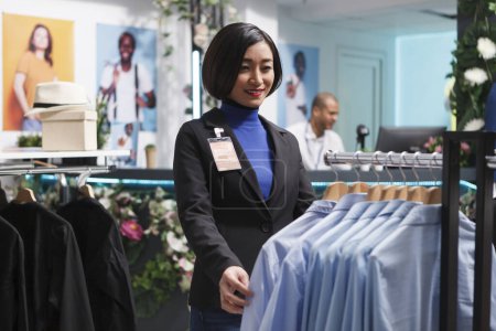 Photo for Clothing store smiling asian woman assistant standing near apparel rack, checking and adjusting merchandise. Shopping mall boutique seller examining formal shirts on display hangers - Royalty Free Image