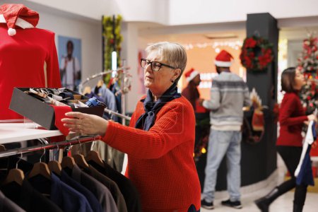 Photo for Client checking accessories box in clothing store, searching for ties and belts to give presents on christmas eve celebration. Elderly woman browsing through clothes on hangers, winter promotions. - Royalty Free Image