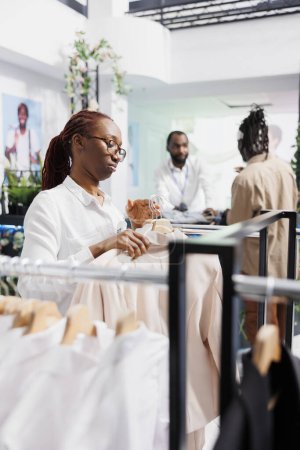 Foto de Asistente de tienda de ropa ajustando expositor para mostrar chaquetas nueva colección para los clientes. Mujer afroamericana arreglando mercancía de ropa de moda en grandes almacenes - Imagen libre de derechos