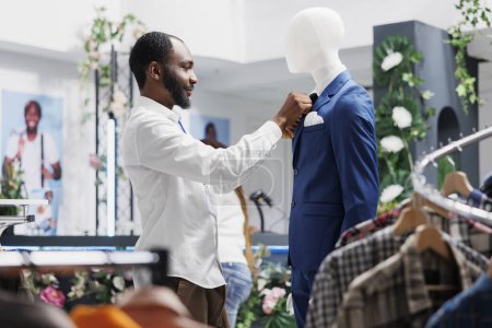 Photo for Clothing store worker fixing tie on mannequin wearing formal suit, showcasing menswear to customers. Fashion boutique african american man adjusting male trendy jacket on display dummy model - Royalty Free Image