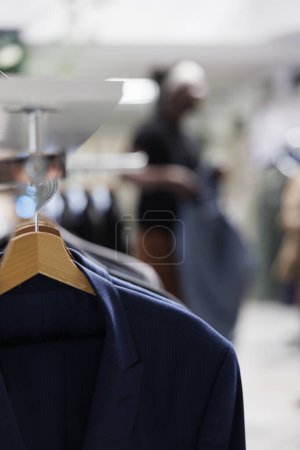 Téléchargez les photos : Chemises décontractées à la mode pour hommes accrochées à un étagère dans un magasin de vêtements avec le client choisissant une tenue sur fond flou. Usure formelle des cintres dans le centre commercial gros plan mise au point sélective - en image libre de droit