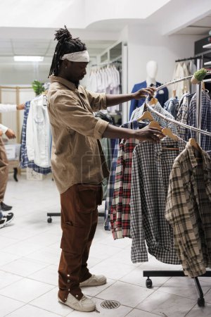 Foto de Hombre afroamericano hojeando ropa en tienda minorista showroom. Joven cliente que comprueba la ropa que cuelga en el estante para comprar camisa a cuadros casual en el centro comercial boutique de moda - Imagen libre de derechos