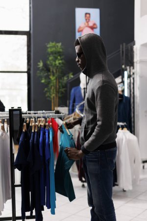 Photo for African american man looking aside while robbing clothing store, checking racks with trendy clothes. Thief trying to steal fashionable merchandise, wearing sunglasses and hood not to be recognized - Royalty Free Image