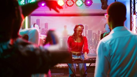 Téléchargez les photos : Femme DJ mixant de la musique dans une boîte de nuit, faisant la fête avec divers groupes de personnes sur la piste de danse. Funky girl s'amuse à la fête disco dans la discothèque, station audio sur scène du club. Coup de main. - en image libre de droit