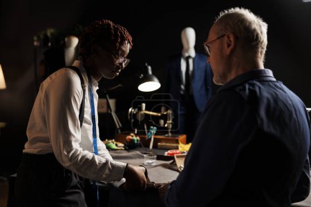 Photo for Attentive african american apprentice carefully listening to master dressmaker guiding her with cutting fabric. Couturiers manufacturing upcoming bespoken fashion collection - Royalty Free Image