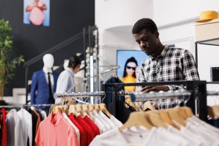 Téléchargez les photos : Homme élégant afro-américain vérifiant chemises décontractées, shopping pour des vêtements à la mode dans la boutique moderne. Client Shopaholic analysant le tissu de marchandises à la mode et le prix dans le magasin de vêtements - en image libre de droit