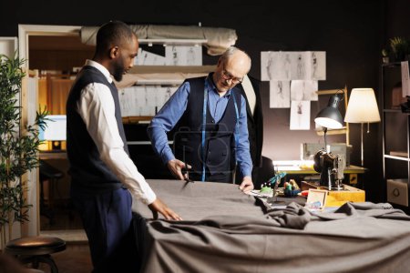 Photo for Meticulous elderly master tailor cutting fabric sheet for upcoming bespoke luxury fashion collection. Senior suitmaker showing african american apprentice expert craftmanship - Royalty Free Image