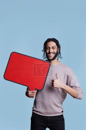 Téléchargez les photos : Homme arabe souriant tenant bannière vierge rouge avec espace de copie et montrant le pouce vers le haut portrait. Personne debout avec bulle de parole vide et geste positif tout en regardant la caméra - en image libre de droit