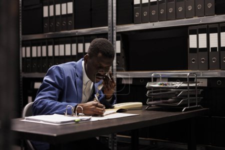 Téléchargez les photos : Le comptable afro-américain discute à distance avec le manager par téléphone fixe, discute du rapport de comptabilité dans la salle de stockage. Homme d'affaires effectuant des heures supplémentaires avec des dossiers administratifs - en image libre de droit