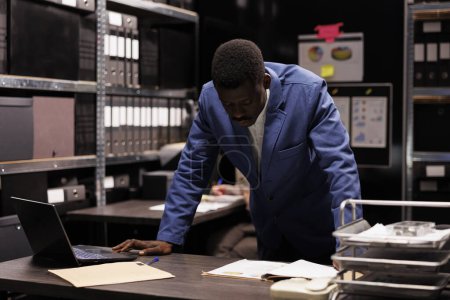 Photo for African american officer analyzing criminal case, checking crime scene evidence in arhive room. Private detective reading criminology report, searching for clues. Law enforcement concept - Royalty Free Image