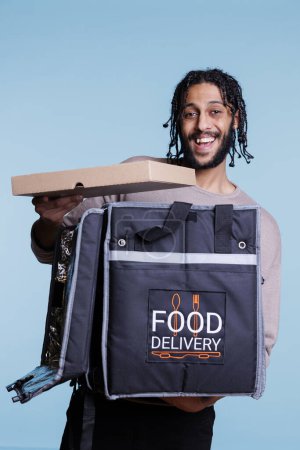 Foto de Feliz mensajero árabe sosteniendo la caja de pizza, entregando comida rápida y mirando a la cámara con expresión alegre. Repartidor de restaurante despreocupado pizzería de pie con retrato de mochila térmica - Imagen libre de derechos