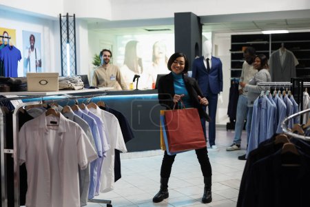 Foto de Mujer asiática sonriente bailando con compras, disfrutando comprando ropa en la tienda de ropa. Boutique alegre cliente celebración de paquetes y hacer movimientos a la música en el centro comercial - Imagen libre de derechos