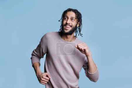 Photo for Cheerful arab man dancing with happy facial expression and looking away with smile. Young adult arabian handsome person in good mood making moves with hands on blue background - Royalty Free Image