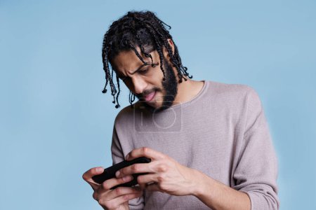 Téléchargez les photos : Focalisé jeune homme arabe tenant téléphone mobile et jouer à un jeu en ligne difficile. Personne avec expression faciale tendue profitant d'un logiciel d'application divertissant sur smartphone - en image libre de droit