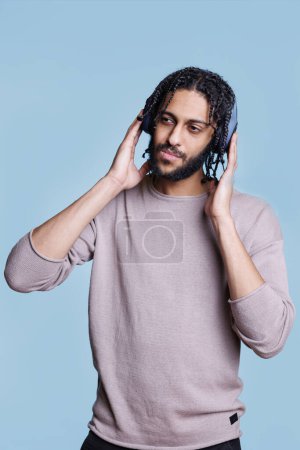 Téléchargez les photos : Arabe écoutant la chanson, touchant écouteurs sans fil sur la tête et détournant les yeux avec expression faciale coûteuse. Personne dans les écouteurs portant des vêtements décontractés appréciant la musique - en image libre de droit