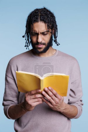 Foto de Estudiante árabe enfocado reflexivo leyendo libro de novela con cubierta amarilla. Hombre adulto joven pensativo concentrado que estudia literatura mientras sostiene libro de texto abierto de tapa blanda - Imagen libre de derechos