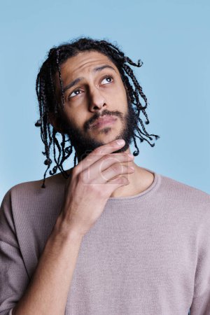 Photo for Arab handsome man thinking and rubbing chin while looking away with puzzled facial expression. Thoughtful attractive model with black braids hairstyle with wondering emotions - Royalty Free Image