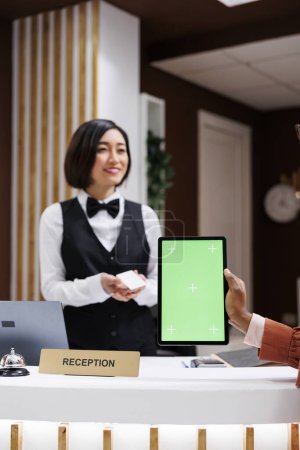 Foto de Mujer sosteniendo tableta con pantalla verde en la recepción, recibiendo la tarjeta de acceso a la habitación de la recepcionista asiática. Joven usando pantalla de maqueta aislada con cromakey en la recepción del hotel. - Imagen libre de derechos