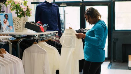 Téléchargez les photos : Client afro-américain magasinant pour chemise blanche, vérifier le tissu des articles dans la boutique moderne. Femme Shopaholic acheter une nouvelle collection de mode, à la recherche de vêtements décontractés dans un magasin de vêtements - en image libre de droit