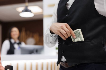 Photo for Hotel staff receives money in lobby, putting cash in pocket to help guests with luggage after check in. Young man working as bellboy accepting cash and tip from tourists. Close up. - Royalty Free Image