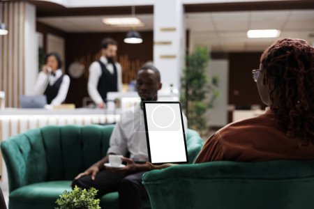 Téléchargez les photos : Les personnes afro-américaines à l'hôtel en utilisant une tablette avec écran blanc, en regardant l'affichage du modèle vierge et assis dans le salon. Dispositif de maintien des invités féminin avec espace de copie isolé. - en image libre de droit