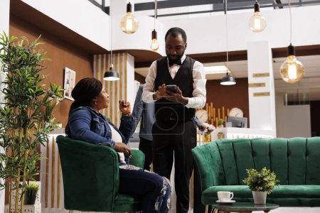 Photo for Young African American woman tourist sitting at luxury hotel lobby making order talking with waiter, ordering food and drinks while waiting for check-in. Customer service in hospitality - Royalty Free Image