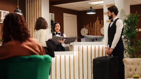 Photo for Tourist arriving in hotel lounge, assisted by helpful bellboy and receptionist managing her reservation details. Cheerful resort personnel maintaining positive attitude while helping customer - Royalty Free Image