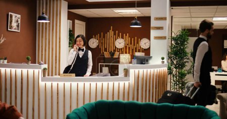 Photo for Happy professional hotel personnel doing various administrative tasks in stylish hotel reception. Asian receptionist taking room booking phone calls and bellboy carrying tourist luggage - Royalty Free Image