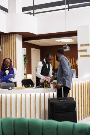 Téléchargez les photos : Le personnel de la réception de l'hôtel afro-américain assiste les clients à la réception, enregistre les touristes, demande des détails de réservation lors de l'enregistrement. Touriste avec bagages arrivant à la chambre de réservation du complexe - en image libre de droit