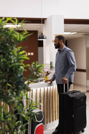 Photo for Young African American man with suitcase standing at reception area, talking pleasantly with front desk employees, waiting in line to check-in after arrival. Black guy tourist checking out of hotel - Royalty Free Image