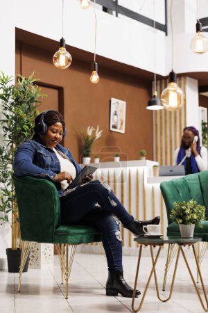 Téléchargez les photos : Femme afro-américaine détendue portant un casque sans fil en utilisant une tablette numérique tout en se relaxant dans une zone de repos confortable de l'hôtel, en regardant un film en ligne et en buvant du café en attendant l'enregistrement - en image libre de droit