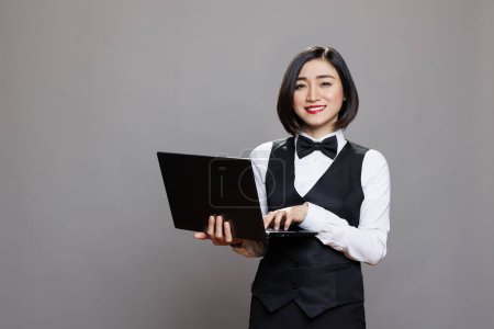 Photo for Smiling asian waitress typing on laptop keyboard and looking at camera with friendly facial expression. Restaurant employee using catering service software on portable computer portrait - Royalty Free Image