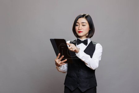 Foto de Mujer asiática recepcionista tocando en la pantalla táctil de la tableta digital mientras trabaja en la industria de la hospitalidad. Servicio de catering camarera en uniforme blanco y negro comprar on-line - Imagen libre de derechos
