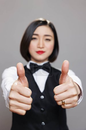 Foto de Mujer asiática alegre en uniforme de camarera sonriendo y dando pulgares hacia arriba símbolo de primer plano. Trabajador de cafetería confirmando y recomendando buen servicio de restaurante y mirando a la cámara - Imagen libre de derechos