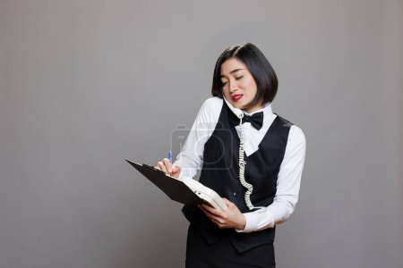Photo for Waitress holding clipboard and talking on landline phone, managing communication and tasks in cafe. Receptionist engaging in conversation on telephone and writing information - Royalty Free Image