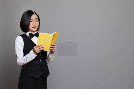 Photo for Concentrated asian woman receptionist reading book while taking break. Catering service young waitress wearing professional uniform learning cafe menu with yellow blank cover - Royalty Free Image