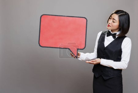 Photo for Young asian waitress holding blank chat balloon mockup while posing in studio. Attractive receptionist in professional uniform showing empty speak cloud with copy space for promo message - Royalty Free Image