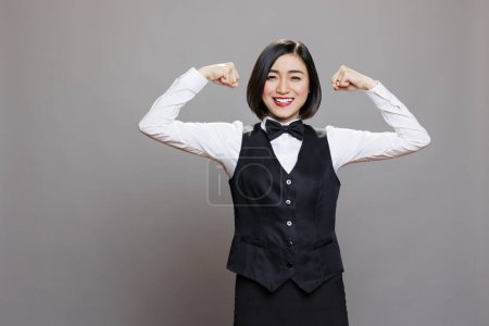 Photo for Smiling asian waitress in uniform showing arms muscles and looking at camera with joyful facial expression. Cheerful receptionist displaying biceps, showcasing strength and power portrait - Royalty Free Image
